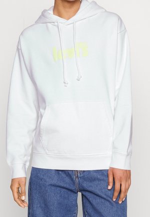 Personne portant un sweat à capuche blanc Levi’s avec poche avant et cordons, associé à un jean bleu, visible des épaules jusqu'aux hanches.