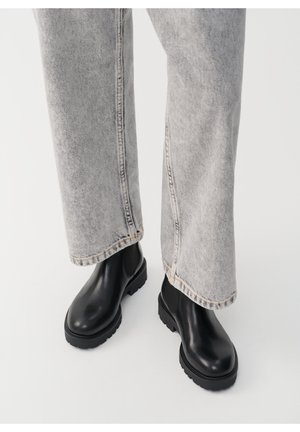 Bottes chevilles en cuir noir avec une semelle épaisse, associées à un pantalon en denim gris clair avec coutures visibles et une légère évasement à l'ourlet.
