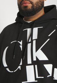 Black hoodie featuring large white graphic text in varying fonts and sizes. Cuffed sleeves with an adjustable drawstring hood.