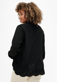Blusa nera con maniche lunghe, caratterizzata da un orlo a scallop con dettagli traforati a forma di fiore. Tessuto leggero con una finitura testurizzata.