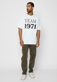 Biała koszulka z grafiką "TEAM 1971" w czerni i złocie, zestawiona z oliwkowo-zielonymi spodniami i białymi trampkami. Luźny krój, krótki rękaw.