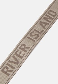 Béžový tkaný pásek s nápisem "RIVER ISLAND" tmavě hnědým písmem, který představuje texturovaný povrch a jednoduchý design.