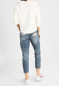 Jean en denim bleu à coupe décontractée, avec broderies de nuages et d'arc-en-ciel sur les poches arrière. Associé à un haut blanc texturé et des baskets.