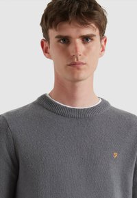 Szary wełniany sweter z prążkowanym okrągłym dekoltem. Posiada małe pomarańczowe logo po lewej stronie. Gładka faktura, dopasowany krój.