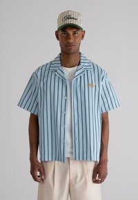 Light blue short-sleeve shirt with vertical navy stripes, featuring a button-up front and an embroidered logo. Paired with cream pants and a cap.