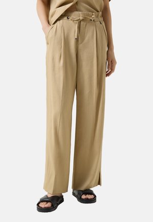 Beige bukser med vide ben, bindebånd i taljen, sidelommer og forreste plisseringer, båret med sorte åbne sandaler, der viser tæerne.