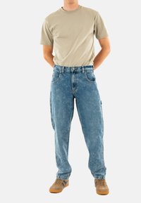Lichtbeige t-shirt met korte mouwen, gecombineerd met een losse, vervaagde blauwe spijkerbroek met voorzakken en een knoopsluiting. Bruine sneakers.