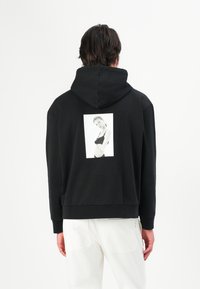 Svart hettegenser laget av mykt stoff, med et rektangulært hvitt grafisk trykk på baksiden. Ledig passform med ribbestrikkede cuffs.