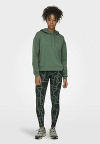 Grüne Kapuzenjacke mit einer Kängurutasche, kombiniert mit schwarzen Leggings, die ein geometrisches Muster in hellgrün aufweisen. Sneakers runden das Outfit ab.