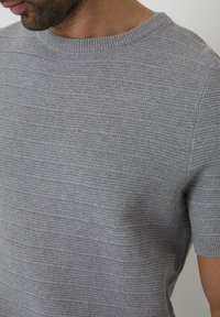 Šedý pletený svetr s krátkým rukávem, zdobený vodorovnými pruhy a ribovaným výstřihem. Textura vypadá mírně vyvýšeně a je jednotná.