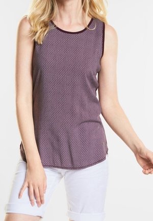Femme portant un haut violet à motifs sans manches et un short blanc jusqu'aux genoux sur un fond uni.