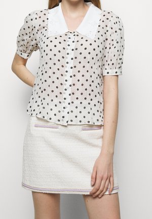 Vrouw met een doorschijnende witte blouse met zwarte polkadots en pofmouwen, gecombineerd met een witte gestructureerde minirok met lavendelkleurige afwerking.