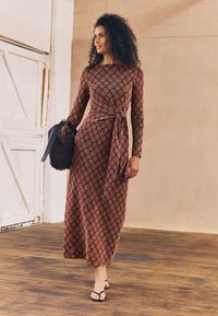 Dlouhé, přiléhavé šaty z vzorované tkaniny s hnědo-červeným motivem. Obsahují dlouhé rukávy a uvázaný pás. Nosené s černými sandály.