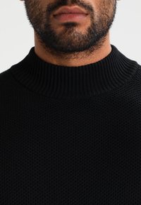 Černý pletený svetr s vysokým žebrovaným límcem, texturovanou tkaninou a těsným střihem kolem krku, který zdůrazňuje jeho minimalistický design.