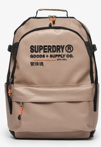 Beige ryggsäck tillverkad av hållbart tyg med svarta dragkedjor, orange dragare och präglad svart logotext. Har flera fack och handtag.