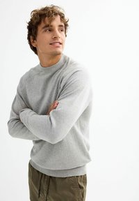 Suéter gris claro con cuello redondo acanalado y puños, combinado con pantalones verde oliva, llevado por un joven con cabello rizado.