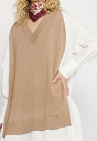 Gilet en maille beige à col en V, en tissu doux, avec des bords côtelés, superposé sur une chemise blanche, avec une écharpe à motifs autour du cou.