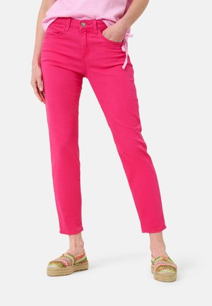 Femme portant un pantalon slim rose vif et des chaussures slip-on tissées colorées, debout avec une main dans la poche.