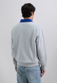 Man met de rug naar de camera, gekleed in een lichtgrijze trui met een blauwe kraag en lichtblauwe denim jeans, tegen een effen achtergrond.