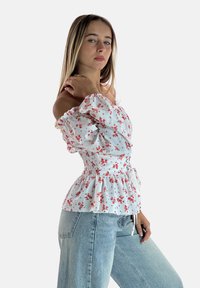 Weiße florale Bluse mit schulterfreien Trägern, smockierter Taille, Rüschenschößen und rosa Akzenten, kombiniert mit lockeren hellblauen Jeans.
