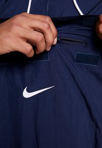 Marinblå vindjacka med en vit Nike-logotyp. Har en dragkedjeficka och kardborreflikar. Materialet har en slät, något skrynklig textur.