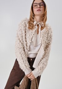 Cardigan crème duveteux avec motif bouclé texturé, manches longues, poignets côtelés, porté sur un chemisier blanc, associé à un pantalon marron foncé.