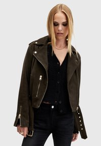 Giacca da biker in suede verde oliva con cerniere argentate, bottoni a scatto e dettagli in borchie; vestibilità corta con tasche a patta e maniche lunghe.