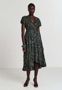 Robe noire avec motif de feuilles vertes et dorées, manches volantées, design enroulé, taille cintrée et ourlet asymétrique. Le mannequin porte des talons noirs et des bracelets.
