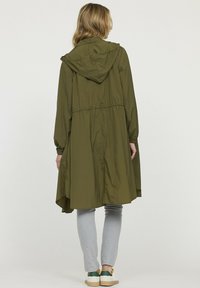 Veste anorak à capuche vert olive, légère avec une taille cintrée, manches longues et un ourlet arrière plus long ; texture lisse et design minimaliste.