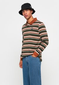 Man met een zwarte bucket hat, groen, bruin en wit gestreepte lange mouwen polo met een bruine kraag, blauwe jeans, poseert met één hand in zijn zak.
