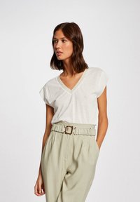 Witte V-hals t-shirt met korte mouwen, gecombineerd met lichtgroene high-waisted broek en een gouden gespriem. Zachte stoftextuur.