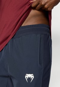 Ναυτικού χρώματος αθλητικό παντελόνι με λεία υφή, ελαστική μέση και λευκό λογότυπο φιδιού στο μηρό, σε συνδυασμό με πουκάμισο βουργουνδί.