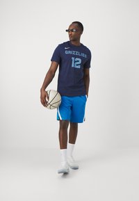 Marineblå basketball t-shirt med "GRIZZLIES 12" i lys blå, parret med blå shorts med hvide side-striber. Holder en basketball.