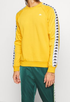 Man draagt een gele sweatshirt met zwarte logoband op de mouwen en groene sportbroek met een witte logostreep langs de zijkant.
