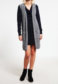 Lång, öppen front cardigan i grått stickat med randiga svarta ärmar. Har två framfickor och ribbade fållar. Bärs över en mörk klänning.