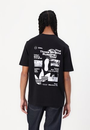 Personne avec de longs cheveux tressés, portant un T-shirt graphique noir avec un texte blanc, des noms de villes et le logo Adidas dans le dos, debout contre un mur blanc.