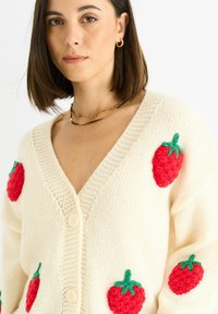 Crèmekleurige gebreide cardigan met rode, textuurlijke aardbei ontwerpen en groene accenten. Heeft een diepe V-hals en twee grote knopen.
