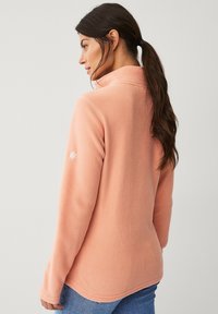 Lichtroze fleece jack met een hoge kraag, lange mouwen en een subtiele logo-accent op de linkermouw. Zachte textuur, aansluitend ontwerp.