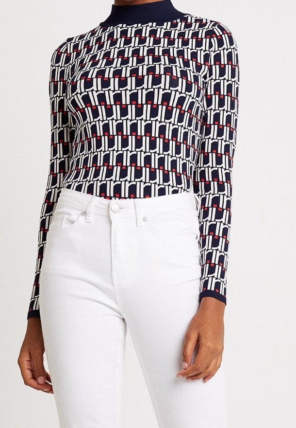 Femme portant un haut à manches longues à motifs bleu marine, blanc et rouge, rentré dans un jean blanc taille haute, debout devant un fond uni.