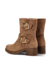 Botas de ante marrón hasta el tobillo con acentos de doble hebilla dorada, punta redondeada y tacón apilado. La textura es suave con un acabado liso.