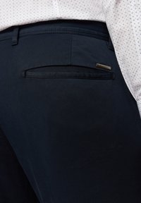 Navyblaue Hose mit einer Gesäßtasche mit Klappendetail und einem kleinen metallischen Etikett. Glattes Gewebe mit subtiler Textur.