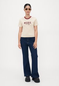 Beige t-shirt with maroon neckline and "MUSE Energy" graphic. Paired with dark blue flared jeans and black platform shoes. Sunglasses worn.