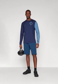 Vyras dėvi tamsiai mėlyną ir šviesiai mėlyną ilgomis rankovėmis dviratininko marškinėlį, mėlynas šortus, juodus sportinius batelius, laiko juodą dviratininko šalmą, stovi ant pilkos grindų.