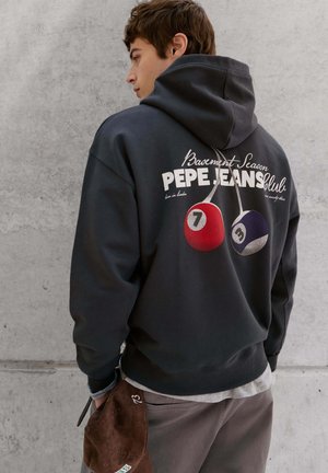 Junger Mann trägt einen dunklen Hoodie mit roten und violetten Billardkugeln und Pepe Jeans-Logo, hält eine braune Kappe und steht vor einer grauen Betonwand.