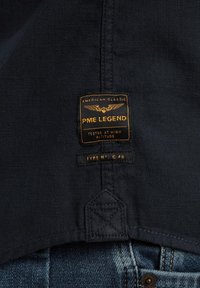 Mornarska tkanina s etiketom z zlatnim tekstom "PME LEGEND", koja sadrži logotip krila i detalje poput "Ispitano na visokoj nadmorskoj visini" i "Tip N° C-46."