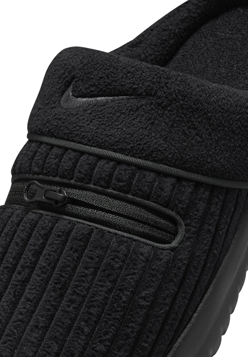 Zapato slip-on de forro polar negro que presenta una superficie texturizada y acanalada, además de un bolsillo con cremallera. El diseño incluye un sutil logotipo en la parte superior.