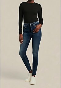 Vaqueros pitillo de lavado oscuro con cintura alta, diseño de cinco bolsillos y un desvanecimiento sutil. Combinados con una camiseta negra de manga larga y zapatillas blancas.