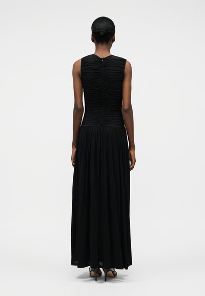 Naine seisab seljaga vaataja poole, kandmas musta varrukateta kleiti, mille ülaosa on voltide ja volditud seelikuosa voogav, kombineeritud mustade kõrgetel kontsadel sandaalidega.