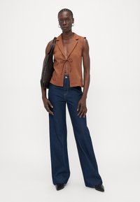 Haut sans manches marron avec une fermeture à nouer, associé à un jean large bleu. Les accessoires comprennent un sac à épaule noir et des boucles d'oreilles créoles.