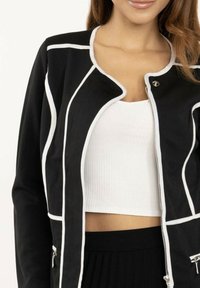 Veste noire avec des finitions blanches, offrant un col arrondi et une fermeture éclair à l'avant. Fabriquée à partir d'un tissu lisse, elle est stylée avec une coupe courte.